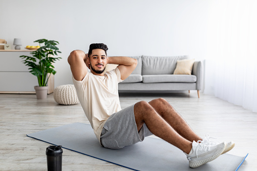Athletic millennial Arab man doing abs exercises, working out at home, full length. Fit Eastern guy doing domestic training during coronavirus epidemic, strengthening core muscles indoors