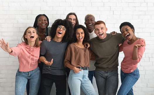Emotional multiracial friends millennials handsome guys and pretty ladies in casual posing together over white brick wall, hugging, cheerfully smiling, laughing at camera. Friendship, friends concept