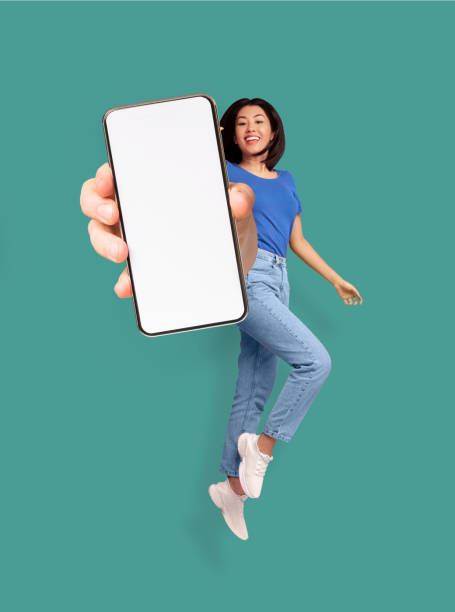 веселая азиатская женщина прыгает и улыбается, показывая мобильный телефон с пустым местом для мобильного приложения или сайта на экране, � - smart phone asian ethnicity mobile phone women стоковые фото и изображения