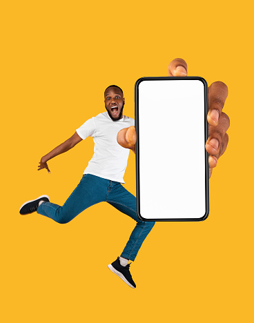 Overjoyed young black man jumping and presenting smartphone with white screen, copy space for mockup ad, website or app design, orange studio background. Creative collage
