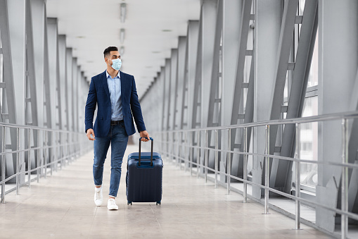 Coronovirus Travels. Arab Businessman Wearing Medical Face Mask Walking With Suitcase At Airport Terminal, Young Middle Eastern Man Carrying Luggage And Going To Departure Gate, Copy Space