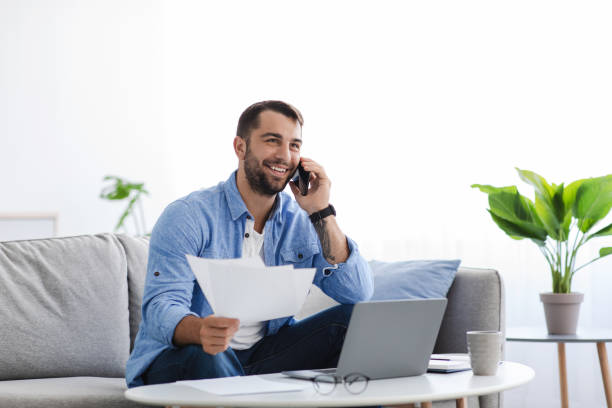 istock Busy smiling adult european guy sitting on sofa work at home with documents, talking with client by phone 1354388453