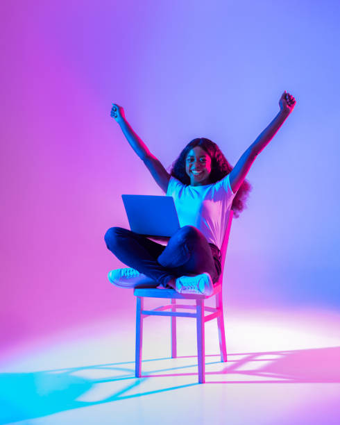 excited young black lady sitting on chair with laptop, raising arms up, celebrating success in neon light - casino worker imagens e fotografias de stock