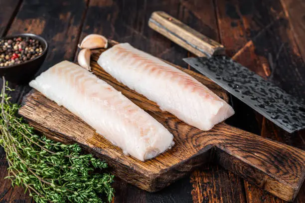 Raw cod loin fillet steak on wooden board with butcher cleaver. Dark wooden background. Top view.