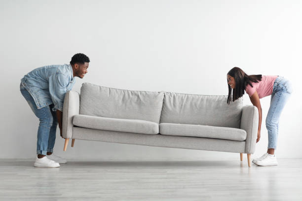 Happy black couple replacing furniture in their new apartment Relocation Concept. Portrait of smiling African American couple moving sofa in new apartment, replacing furniture at home, happy black man and woman placing sofa in cozy studio flat, profile side view real wife stories stock pictures, royalty-free photos & images
