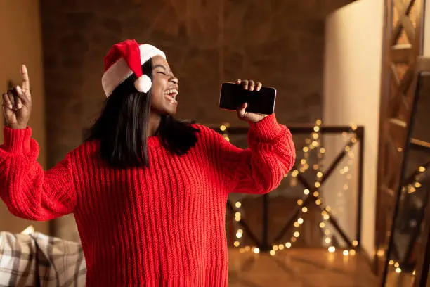 Photo of Christmas holiday music. Happy black lady in Santa hat singing, using smartphone as mic at home, mockup for mobile app