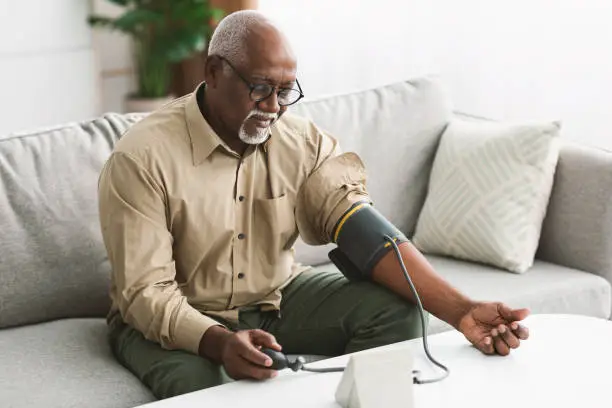 Senior African American Male Measuring Arterial Blood Pressure Having Hypertension Symptom Sitting On Couch At Home. High Blood-Pressure, Health Problem Concept