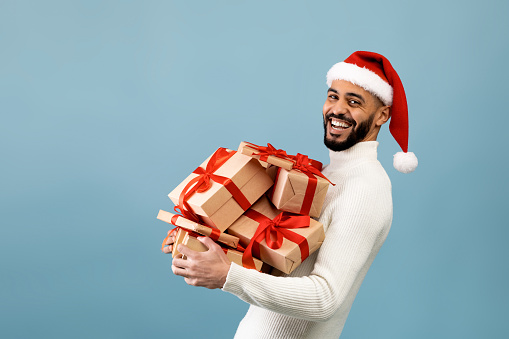 Time for celebration. Overjoyed arab man in santa hat holding stack of gift boxes and smiling at camera, posing over blue background, free space