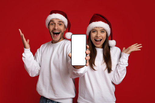 Amazing Offer. Excited Couple In Santa Hats Demonstrating Smartphone With Blank White Screen, Emotional Young Man And Woman Recommending Mobile App Or Website For Online Cristmas Shopping, Mockup