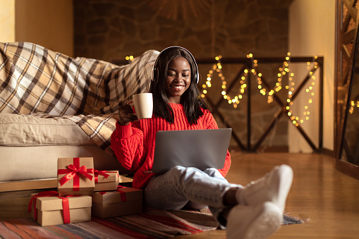 Smiling African American woman celebrating quiet Christmas alone, using laptop, watching Xmas movie, drinking coffee near gift boxes at home. Winter holidays, New Year eve concept