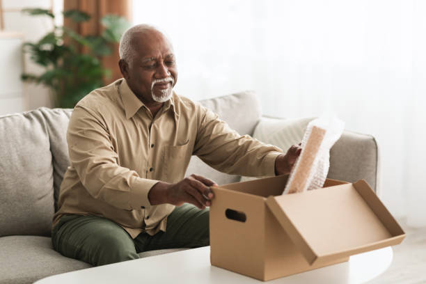 Retired Senior African American Man Packing Stuff In Box Indoors Retirement Concept. Senior African American Man Packing Stuff Putting His Belongings In Cardboard Box Sitting On Sofa Indoors. Retired Lifestyle And End Of Career Concept belongings stock pictures, royalty-free photos & images