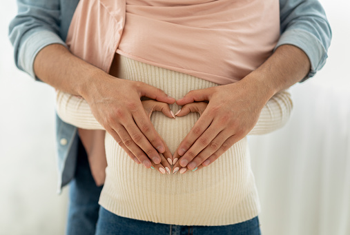 Closeup view of muslim pregnant woman and her husband hugging tummy together, making heart with their hands. Romantic arab couple expressing love to their future baby