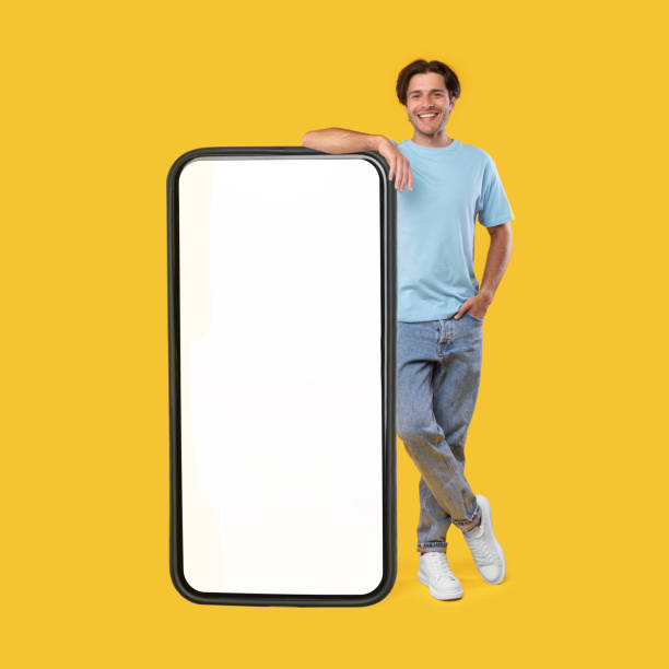 Man leaning on big white empty smartphone screen Full body length of smiling casual guy standing and leaning on huge giant cell phone with empty white screen template, orange studio background. Mock up for app website, free space for advertising leaning stock pictures, royalty-free photos & images