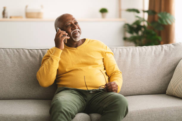 uomo afroamericano anziano che parla al cellulare seduto a casa - senior adult african descent men black foto e immagini stock