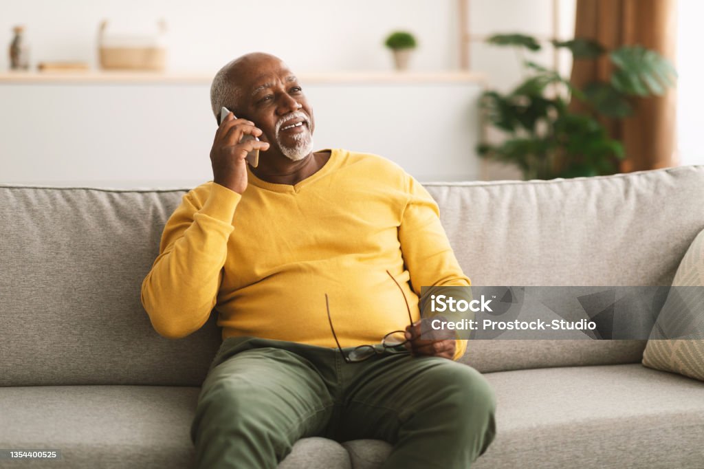 Senior African American Man Talking On Cellphone Sitting At Home Senior African American Man Talking On Cellphone Having Pleasant Phone Conversation Sitting On Sofa At Home, Looking Aside And Smiling. Mobile Communication Concept Using Phone Stock Photo