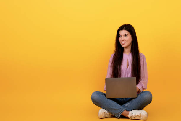 istock Happy armenian woman using laptop computer on yellow studio background, sitting on floor and looking at copy space 1354400756