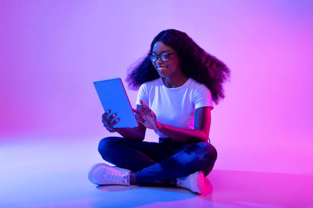 Full length of young black lady using tablet pc, studying or working remotely, having online conference in neon light Full length of young black lady using tablet pc, studying or working remotely, having online conference in neon light. Millennial woman with touch pad checking new app, shopping on web fluorescent photos stock pictures, royalty-free photos & images