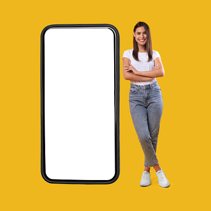 Full body length of smiling casual lady standing with folded arms near giant mobile phone, empty white screen template, orange studio background. Mock up for app or website, free space for advertising