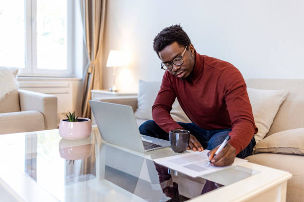 African-American male student or worker in glasses is watching online lectures or webinars and writing notes in a notebook.