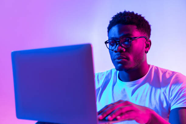 Focused young black man in glasses using laptop computer for online work or communication in neon light Focused young black man in glasses using laptop computer for online work or communication in neon light. Millennial African American guy having distance meeting, video conferencing on web fluorescent photos stock pictures, royalty-free photos & images