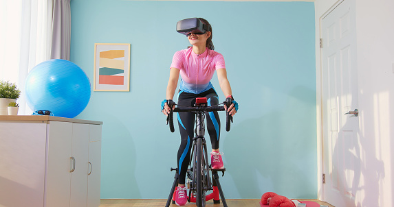 Woman cycling with VR glasses in her room - Asian female cyclist is playing online bike games