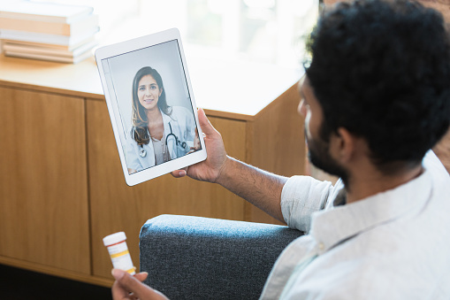 A young male patient discusses a prescription medication with a female doctor via video conference. The man is using a digital tablet to communicate with the doctor.