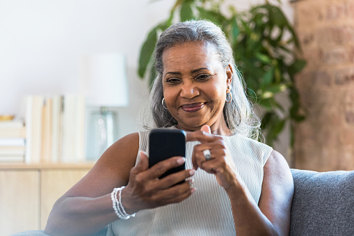 A beautiful senior woman scrolls through social media on her smartphone while relaxing in her home.