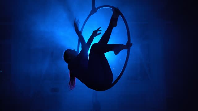 Silhouette, aerial gymnast performs trick in ring in smoky room with backlit
