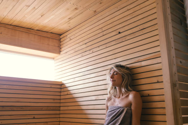 Spa Day: Beautiful Young Blonde Woman Sitting in Sauna Wrapped in a Grey Towel A young Caucasian woman wrapped in a gray towel sitting in a sauna. sauna stock pictures, royalty-free photos & images