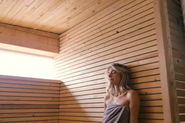A young Caucasian woman wrapped in a gray towel sitting in a sauna.