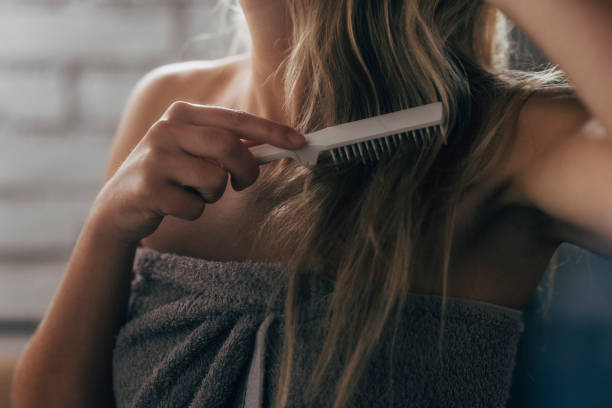 Anonymous Caucasian Woman Combing her Long Blond Hair after Taking a Shower An unrecognizable blonde woman standing in the bathroom wrapped in a gray towel, combing her hair. woman wrapped in bath towel stock pictures, royalty-free photos & images