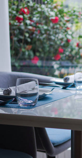 istock Drinking glass, cyan table mat and tableware on white table with flowering plants near window 1354419701