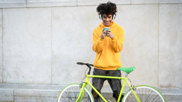 Happy African man using mobile smartphone outdoor while riding with bike in the city - Youth millennial generation lifestyle and technology concept stock photo