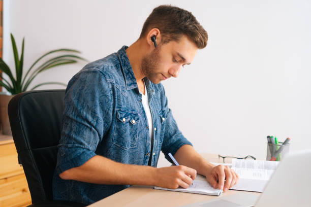 istock Side view of concentrated male student studying online from home using laptop writing notes, watching video class lesson, listening audio course. 1354421793
