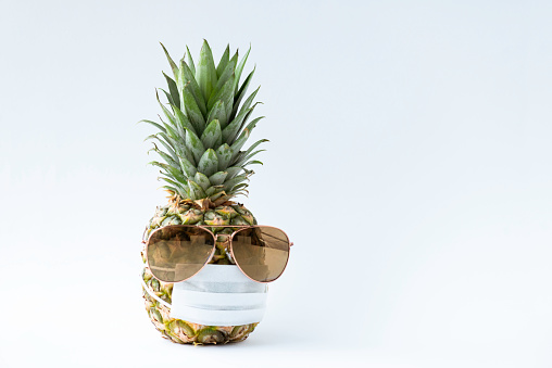 Pineapple wearing sunglasses and a protective face mask in front of white background. Representing virus situations in holiday season.