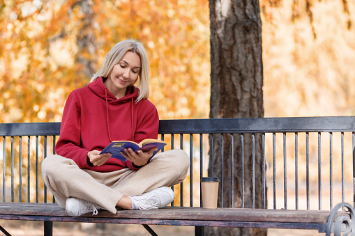Digital detox. Adult lovely woman in casual wear sits on a bench and reading a book in the autumn urban park outdoor, selected focus. Independence, silence, education, privacy, intelligence, leisure concept