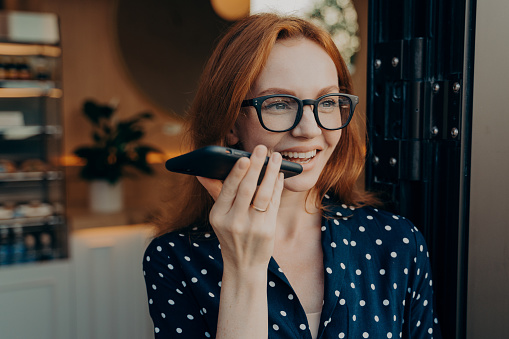 Portrait of happy smiling business woman in spectacles talks on cell phone on loudspeaker while leaving coffee shop in morning, redhead female record audio message while spending time outdoors