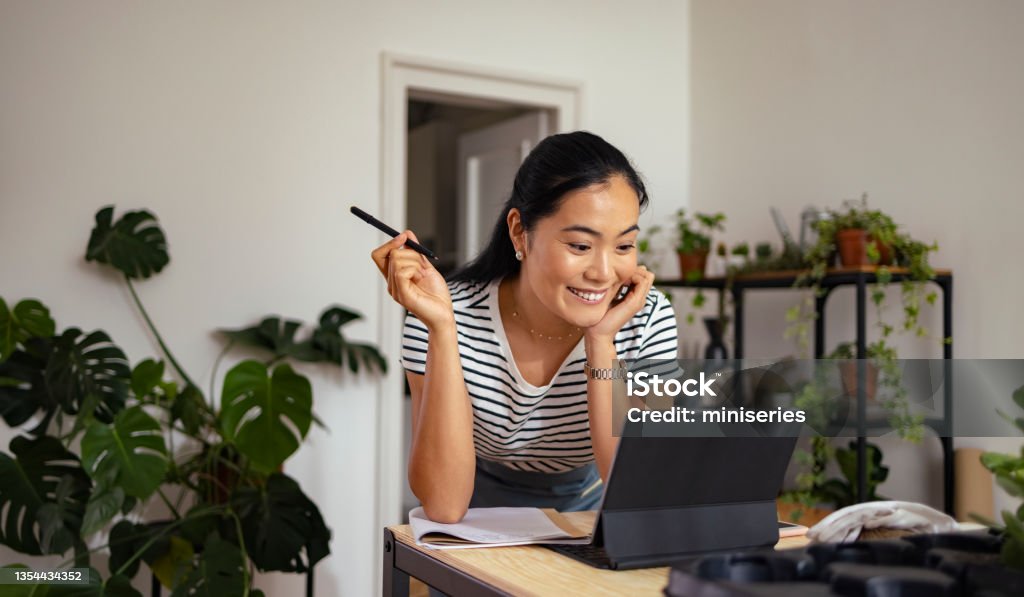 Cheerful Woman Gardener Making Video Call on a Digital Tablet in her Flower Shop Smiling Asian woman talking with client on online video call meeting while standing in her workshop. Business Stock Photo