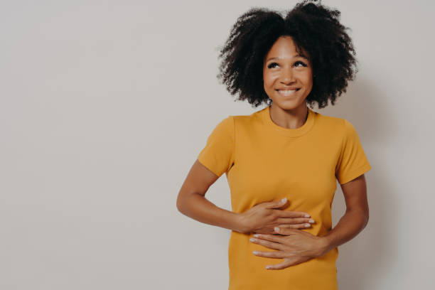 Happy african american woman laughing out loud at some hilarious joke, keeping hands on stomach Happy young african american woman laughing out loud at some hilarious funny joke from friend, keeping both hands on stomach, looking aside cheerfully while standing isolated over white background digestive stock pictures, royalty-free photos & images