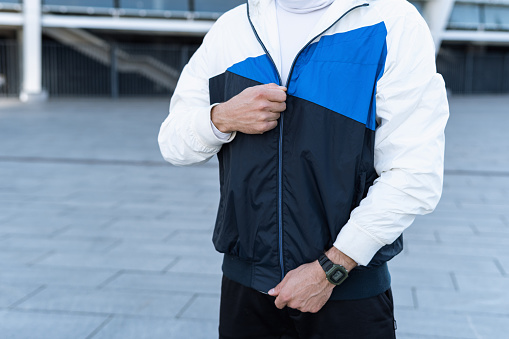 Advertising concept for running clothes. Cropped view of sportive man with smartwatch zips up his sportswear jacket, standing against urban background, prepare to sport training outdoors