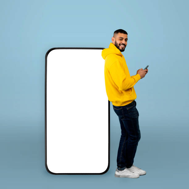 istock Great app. Happy arab guy leaning on big smartphone with blank screen and using his cellphone, blue background, mockup 1354463274