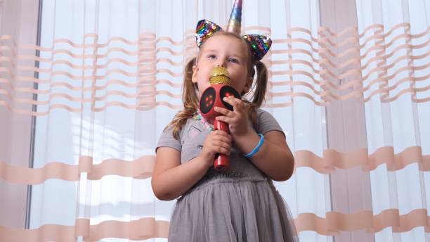 забавная милая маленькая девочка 4-5 лет, поющая в караоке-микрофон, с повязкой единорога, ребенок поет караоке-музыку, веселится на мероприя - cute 4 5 years little girls party стоковые фото и изображения