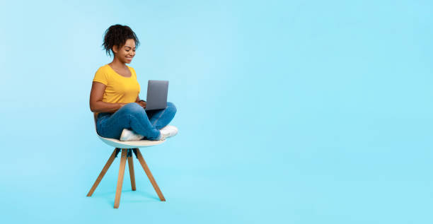 feliz dama negra sentada en la silla, usando la computadora portátil, estudiando en línea, viendo seminarios web o teniendo reuniones remotas, espacio libre - red chairs fotografías e imágenes de stock