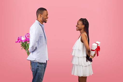 Celebration Concept. Profile side view of excited African American couple making surprise for each other, guy and lady hiding presents behind back, greeting with anniversary or St Valentine's Day