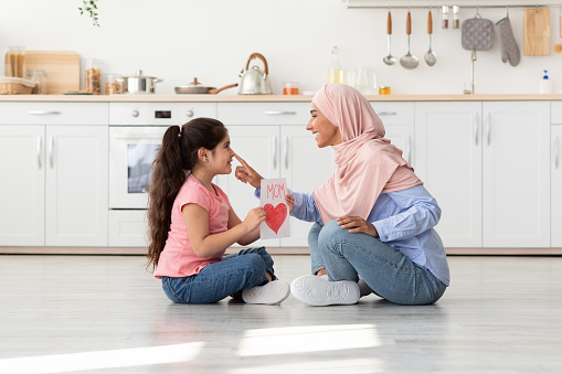 Mother's Day Concept. Portrait Of Loving Little Girl Presenting Handmade Card To Her Muslim Mom In Hijab, Happy Islamic Mommy With Cute Female Child Sitting On Floor in Kitchen, Bonding At Home