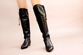 Caucasian female legs in sexy black leather high long boots jackboots on light pink beige background. Copy space in right side, front view. Studio shot photo, closeup.