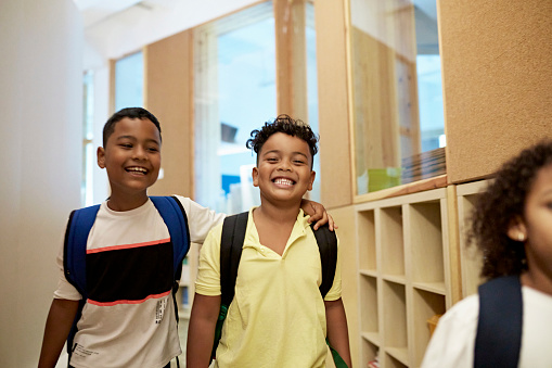 Waist-up view of affectionate children wearing casual clothing, backpacks, and laughing as they look at camera in hallway on the way to class.