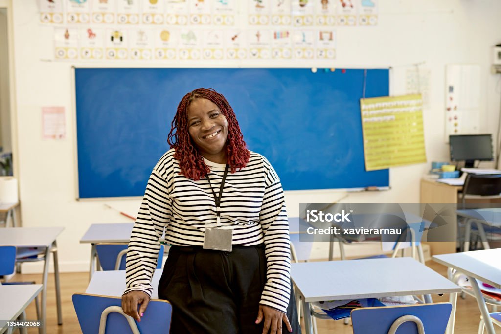 Classroom Portrait of Cheerful School Teacher Front view of mature black woman with curly dyed hair wearing casual clothing and sitting on student desk smiling at camera. Teacher Stock Photo