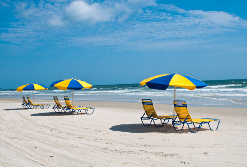 Three beach chairs and an umbrella under a sky with fair weather clouds await sunbathers on the hard packed sand of Daytona Beach, Florida