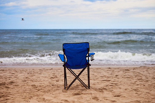 Blue folding chair back on seashore, sea background. Blue chair on sea beach, without people. Beach holiday alone, nobody on Baltic Sea beach, loneliness and hopelessness